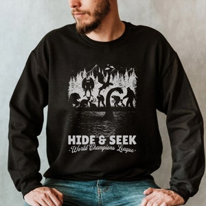 May include: Black sweatshirt featuring a graphic print of various mythical creatures playing hide and seek in a forest. The text "HIDE & SEEK World Champions League." is printed below the graphic.