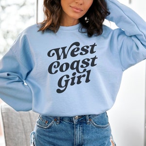May include: A light blue sweatshirt with the text "West Coast Girl" in a bold, black, retro font. The sweatshirt is a crew neck style and appears to be made of a soft material. The overall aesthetic is casual and comfortable.