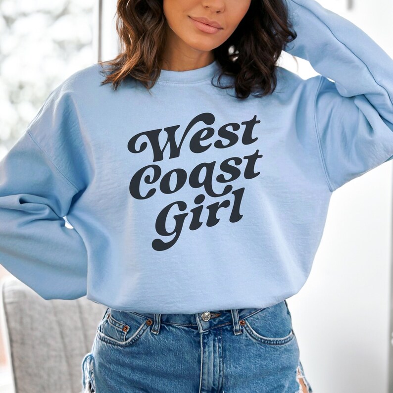 May include: A light blue sweatshirt with the text "West Coast Girl" in a bold, black, retro font. The sweatshirt is a crew neck style and appears to be made of a soft material. The overall aesthetic is casual and comfortable.