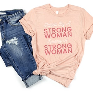 May include: A peach-coloured t-shirt with the text "Raised By A Strong Woman Raising A Strong Woman" in pink lettering.