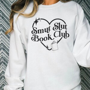 以下が含まれることがあります： 「Smut Slut Book Club」と書かれた黒いハート型のデザインが施された白いスウェットシャツ。デザインには、開いた本と星のアクセントが含まれています。カジュアルで快適なスタイルです。