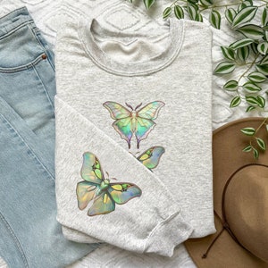 May include: A gray sweatshirt with a design of three iridescent butterflies. The butterflies are green, blue, and purple.