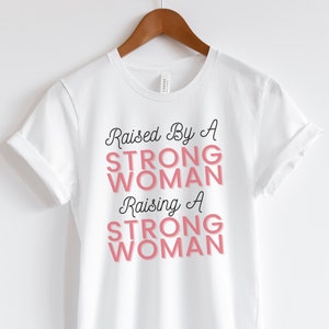 Puede incluir: Camiseta blanca con el texto "Raised by a Strong Woman Raising a Strong Woman" en letras negras y rosas.