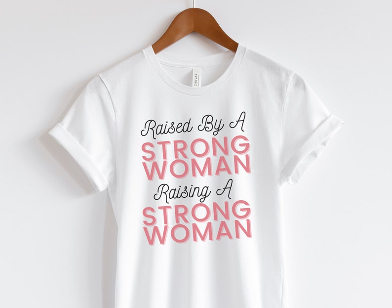 Puede incluir: Camiseta blanca con el texto "Raised by a Strong Woman Raising a Strong Woman" en letras negras y rosas.