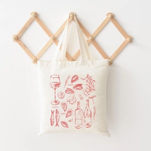 Op de afbeelding: Een witte canvas draagtas met een rode illustratie van wijn, zeevruchten en andere etenswaren. De illustratie bevat een glas wijn, twee flessen wijn, oesters, mosselen, sardines en druiven. De tekst op de tas is "mosselen", "oesters" en "sardientjes".