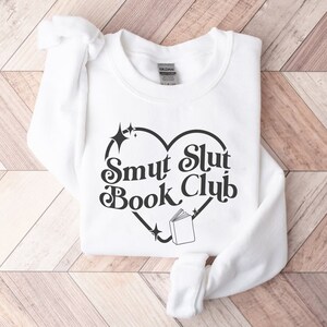 以下が含まれることがあります： ハート型のデザインの中に「Smut Slut Book Club」の文字が入った白いスウェットシャツ。星と本のイラストが描かれています。折りたたまれたスウェットシャツは、袖と裾が見えています。