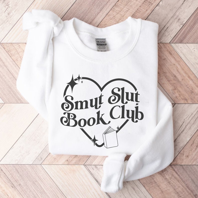 以下が含まれることがあります： ハート型のデザインの中に「Smut Slut Book Club」の文字が入った白いスウェットシャツ。星と本のイラストが描かれています。折りたたまれたスウェットシャツは、袖と裾が見えています。