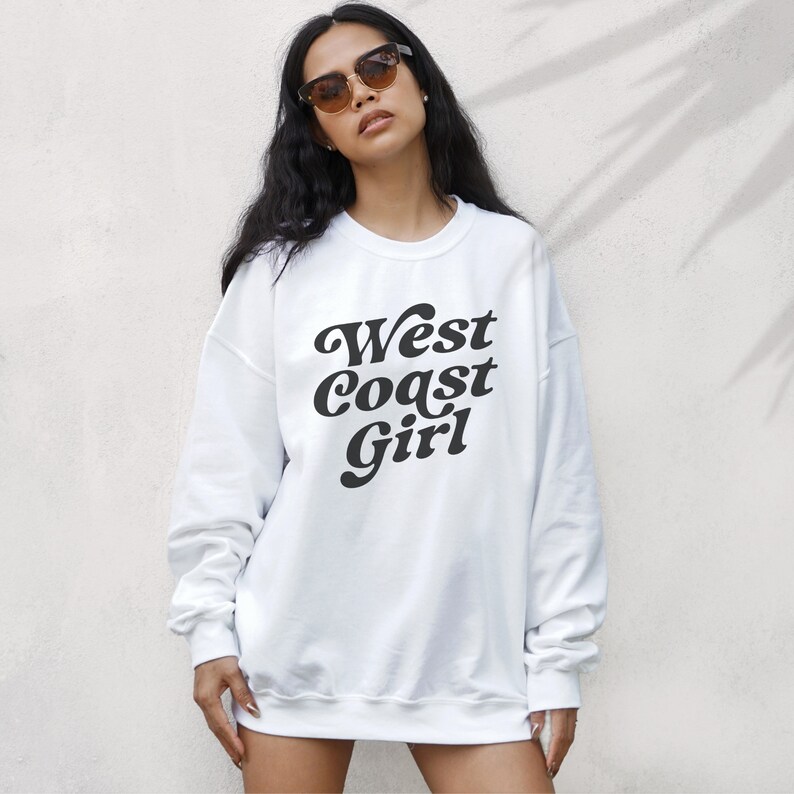 May include: White sweatshirt with the text "West Coast Girl" in a black, retro font. The long-sleeved top is made of a soft material and is suitable for casual wear. The sweatshirt is a classic, comfortable style.
