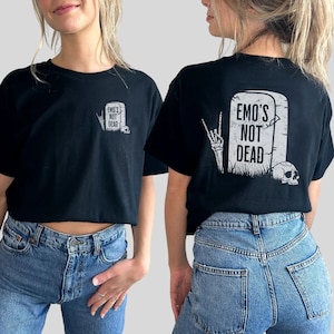 May include: Black cropped t-shirt with a white graphic of a tombstone that reads "Emo's Not Dead" and a skeleton hand making the rock and roll sign. The t-shirt is being modeled by a person wearing blue jeans.