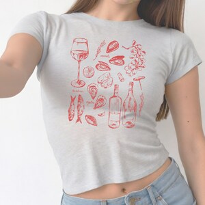 May include: A grey t-shirt with a red graphic design of various seafood, including oysters, mussels, sardines, and wine. The text "oysters", "mussels", and "sardines" is included in the design.