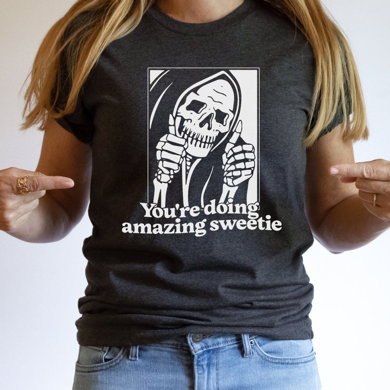 May include: A dark grey t-shirt featuring a white graphic of a hooded skeleton giving a thumbs up with each hand. The text "You're doing amazing sweetie" is printed below the graphic.