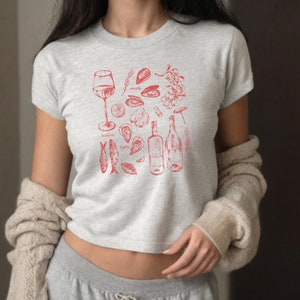 May include: A grey cropped t-shirt with a red illustration of wine, oysters, mussels, and sardines. The text "oysters", "mussels", and "sardines" is included in the design.