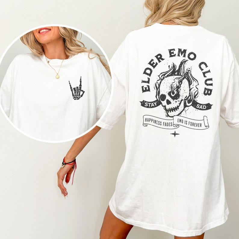 May include: White t-shirt with a black skull graphic on the back. The graphic features a skull with flames and the text "Elder Emo Club". The text "Stay Sad" is above the skull and "Happiness Fades Emo is Forever" is below the skull. The front of the shirt has a black skeleton hand making a rock and roll sign.