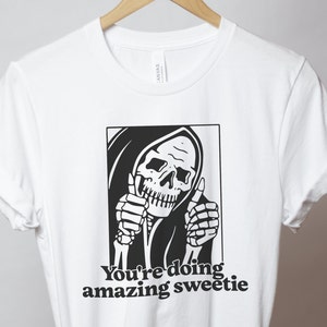 May include: White t-shirt featuring a black and white illustration of the Grim Reaper giving a thumbs up. The text "You're doing amazing sweetie" is printed below the illustration.