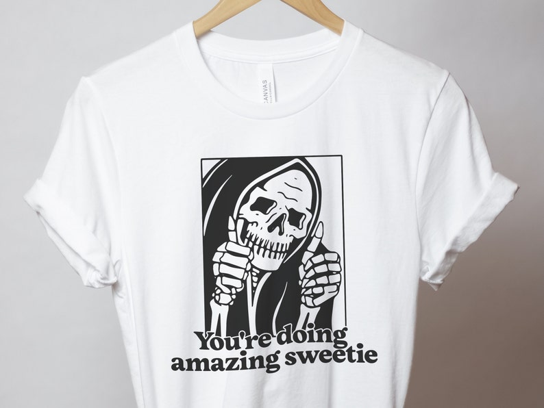 May include: White t-shirt featuring a black and white illustration of the Grim Reaper giving a thumbs up. The text "You're doing amazing sweetie" is printed below the illustration.