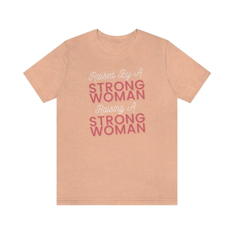 Puede incluir: Una camiseta de color melocot&oacute;n con el texto "Raised By A Strong Woman Raising A Strong Woman" en letras blancas y rosas.