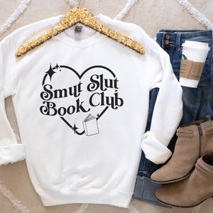 以下が含まれることがあります： ハート型のデザインの中に「Smut Slut Book Club」の文字が入った白いスウェットシャツ。デザインには本と星が含まれています。スウェットシャツは、ブルージーンズ、コーヒーカップ、タンカラーのアンクルブーツと一緒に表示されています。