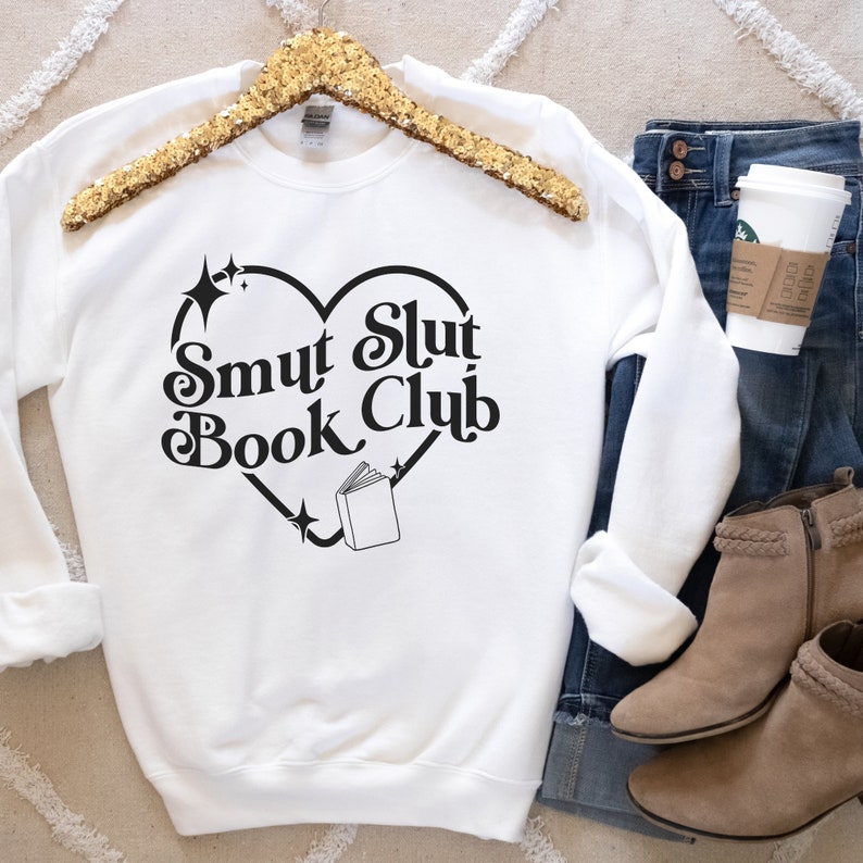 以下が含まれることがあります： ハート型のデザインの中に「Smut Slut Book Club」の文字が入った白いスウェットシャツ。デザインには本と星が含まれています。スウェットシャツは、ブルージーンズ、コーヒーカップ、タンカラーのアンクルブーツと一緒に表示されています。