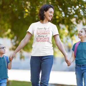 Puede incluir: Una mujer con una camiseta blanca con el texto "Raised By A Strong Woman Raising A Strong Woman" en letras rosas camina con dos ni&ntilde;as peque&ntilde;as. Las ni&ntilde;as van de la mano de la mujer y llevan pantalones vaqueros azules y camisas de color turquesa.