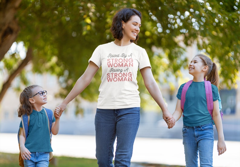 Puede incluir: Una mujer con una camiseta blanca con el texto "Raised By A Strong Woman Raising A Strong Woman" en letras rosas camina con dos ni&ntilde;as peque&ntilde;as. Las ni&ntilde;as van de la mano de la mujer y llevan pantalones vaqueros azules y camisas de color turquesa.