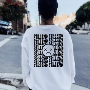 May include: White sweatshirt with a black graphic print of a sad face with an X over the eyes and the text "STILL EMO" repeated multiple times.