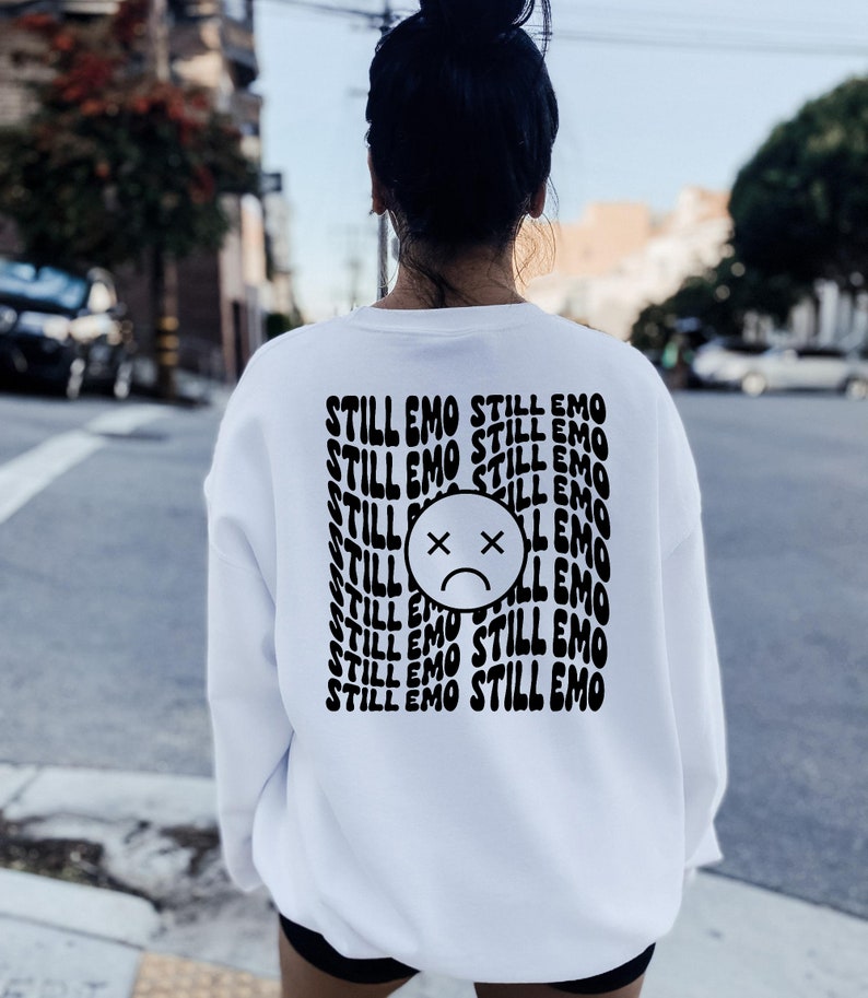 May include: White sweatshirt with a black graphic print of a sad face with an X over the eyes and the text "STILL EMO" repeated multiple times.