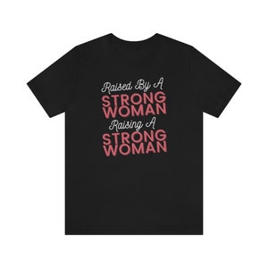 Puede incluir: Camiseta negra con el texto "Raised By A Strong Woman Raising A Strong Woman" en letras blancas y rosas.
