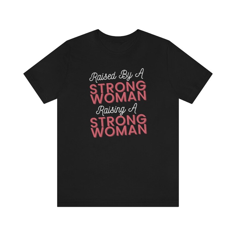 Puede incluir: Camiseta negra con el texto "Raised By A Strong Woman Raising A Strong Woman" en letras blancas y rosas.