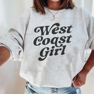 May include: A light gray sweatshirt with the text "West Coast Girl" in a bold, black, retro font. The sweatshirt has long sleeves and a crew neck. The overall style is casual and comfortable.