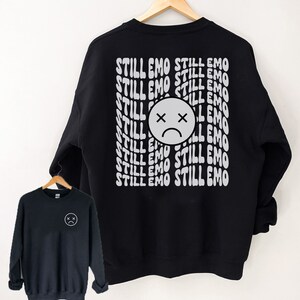 May include: Black sweatshirt with a white graphic print of a sad face and the text "STILL EMO" repeated multiple times.