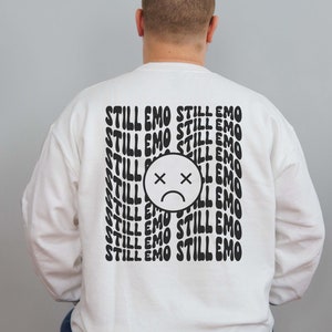 May include: White sweatshirt with black text that reads "STILL EMO" repeated multiple times. A black and white sad face with an X over the eyes is centered on the sweatshirt.