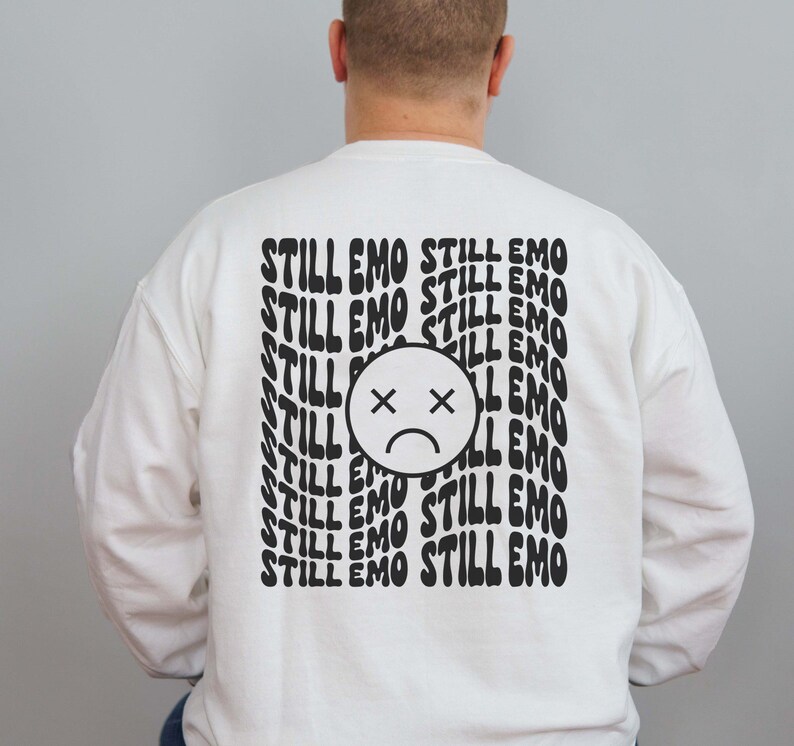 May include: White sweatshirt with black text that reads "STILL EMO" repeated multiple times. A black and white sad face with an X over the eyes is centered on the sweatshirt.