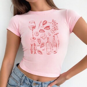 May include: Pink cropped t-shirt with a red graphic print of various seafood, wine, and a glass of wine. The graphic includes the words "mussels", "oysters", "sardines", and "oyster".