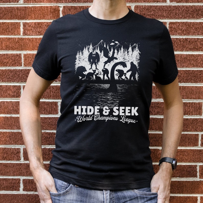 May include: Black t-shirt with a white graphic of various mythical creatures playing hide and seek in a forest. The text "Hide & Seek. World Champions League." is printed below the graphic.