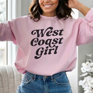May include: A light pink sweatshirt with the text "West Coast Girl" in a black, retro font. The sweatshirt is a crew neck style and is paired with blue jeans. The overall aesthetic is casual and comfortable.