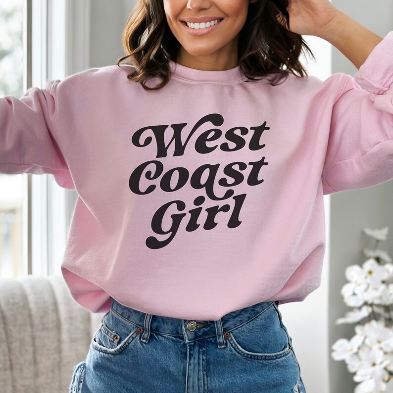 May include: A light pink sweatshirt with the text "West Coast Girl" in a black, retro font. The sweatshirt is a crew neck style and is paired with blue jeans. The overall aesthetic is casual and comfortable.
