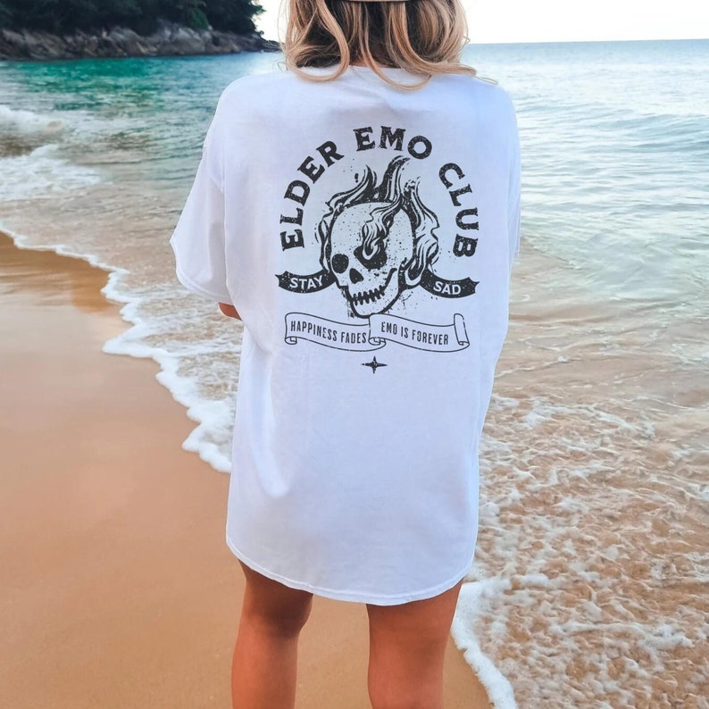 May include: White t-shirt with a black graphic of a skull with flames and the text "Elder Emo Club" with the text "Stay Sad" and "Happiness Fades Emo is Forever" on a banner.