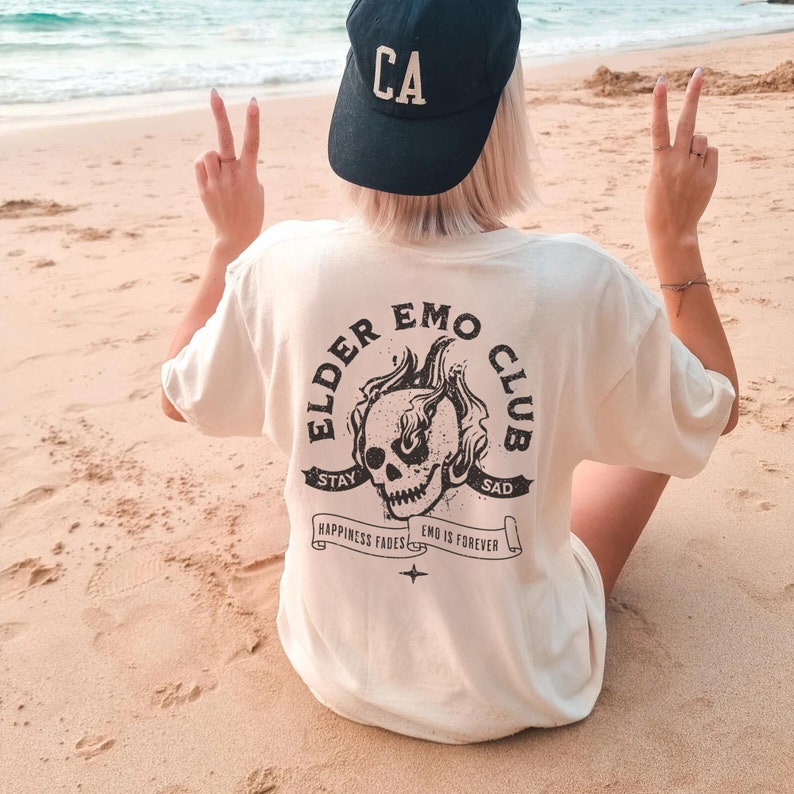 May include: A white t-shirt with a black graphic of a skull with flames on it. The graphic is surrounded by a circle with the text "Elder Emo Club" and the words "Stay Sad" and "Happiness Fades Emo Is Forever".