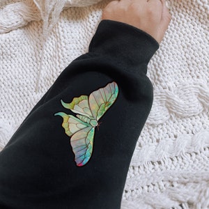 May include: Black sweatshirt with a colorful butterfly graphic print. The butterfly has iridescent wings with shades of green, blue, and purple.
