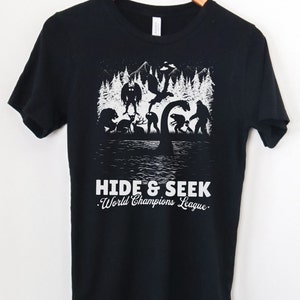 May include: Black t-shirt with a white graphic of various mythical creatures in a forest setting. The text "HIDE & SEEK. World Champions League." is printed below the graphic.