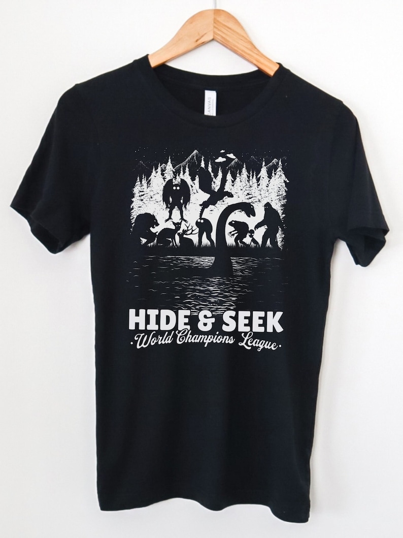 May include: Black t-shirt with a white graphic of various mythical creatures in a forest setting. The text "HIDE & SEEK. World Champions League." is printed below the graphic.