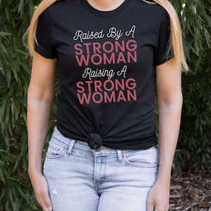 Puede incluir: Una camiseta negra con el texto "Raised By A STRONG WOMAN Raising A STRONG WOMAN" en letras blancas y rosas.