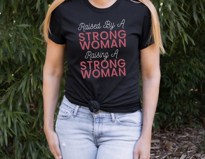Puede incluir: Una camiseta negra con el texto "Raised By A STRONG WOMAN Raising A STRONG WOMAN" en letras blancas y rosas.