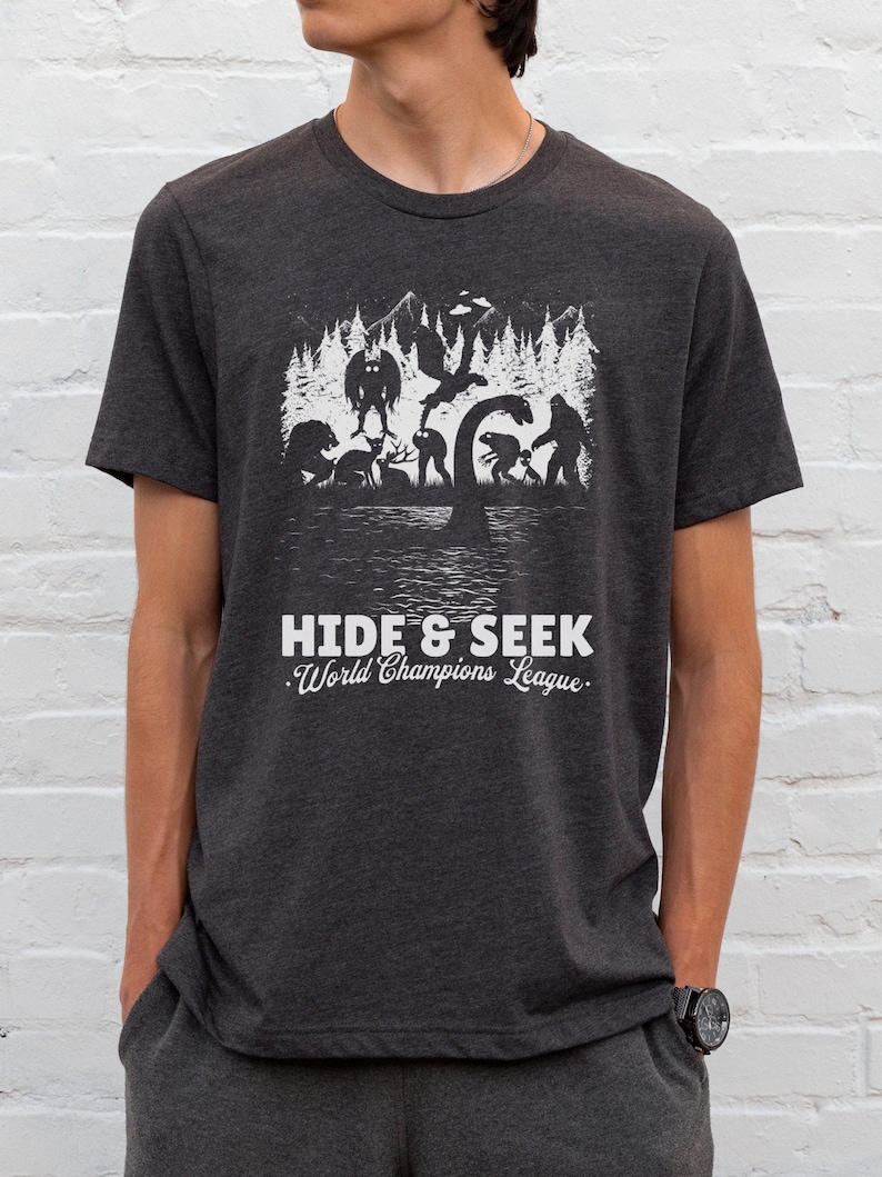 May include: A dark heather grey t-shirt with a white graphic print of various mythical creatures playing hide and seek in a forest. The text "Hide & Seek World Champions League" is printed below the graphic.