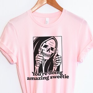 May include: Pink t-shirt featuring a black and white graphic of the Grim Reaper giving a thumbs up. The text "You're doing amazing sweetie" is printed below the graphic.