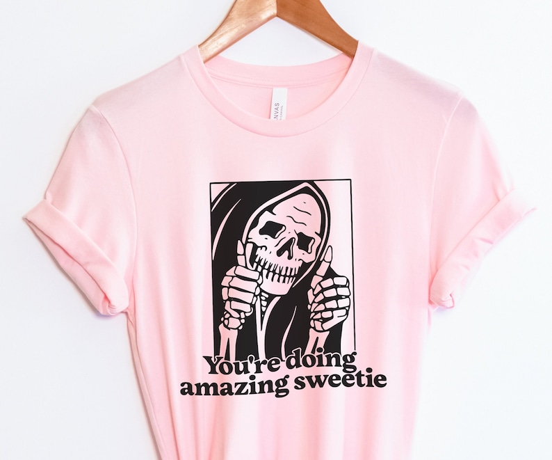 May include: Pink t-shirt featuring a black and white graphic of the Grim Reaper giving a thumbs up. The text "You're doing amazing sweetie" is printed below the graphic.