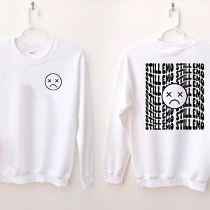 May include: White long-sleeve sweatshirt with a black sad face on the front and the text "STILL EMO" repeated on the back.