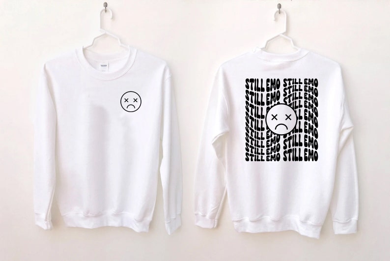 May include: White long-sleeve sweatshirt with a black sad face on the front and the text "STILL EMO" repeated on the back.