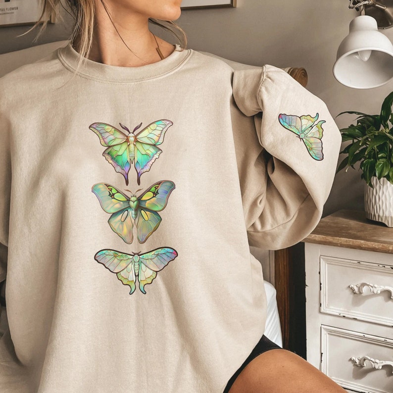 May include: A beige sweatshirt with a graphic print of four iridescent butterflies. The butterflies are arranged vertically, with the largest butterfly at the top and the smallest butterfly at the bottom.