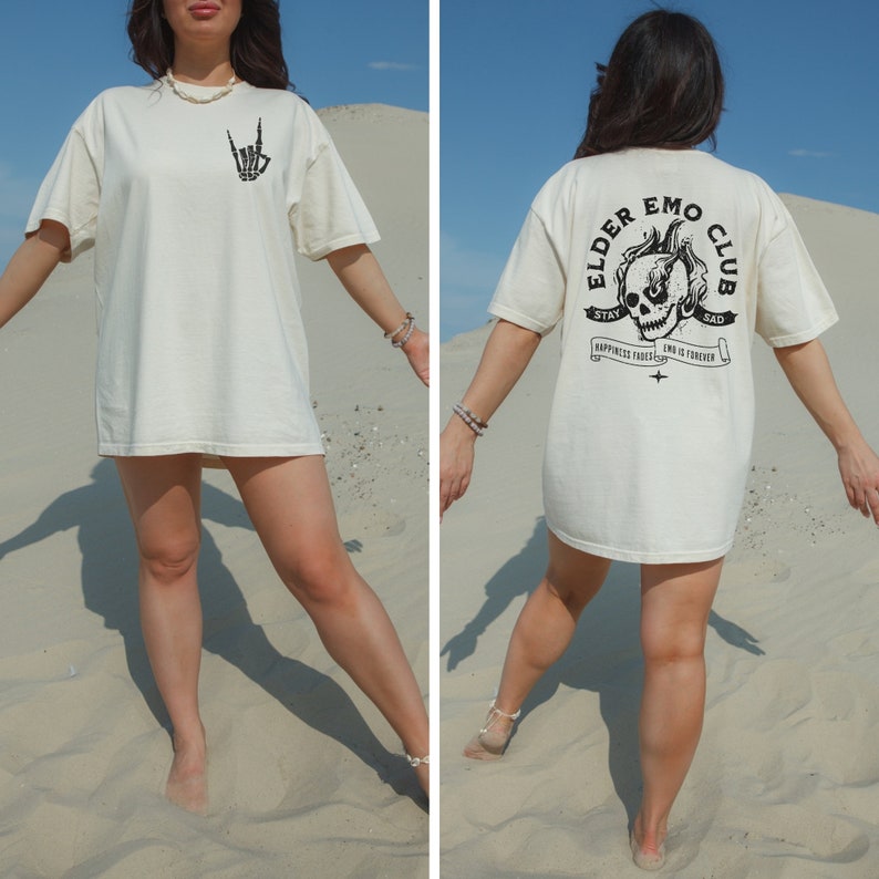 May include: A white oversized t-shirt with a black graphic on the front of a skeleton hand making a rock and roll sign. The back of the shirt has a black graphic of a skull with flames and the text "Elder Emo Club" with the words "Stay Sad" above the skull and "Happiness Ends, Sad Is Forever" below the skull.
